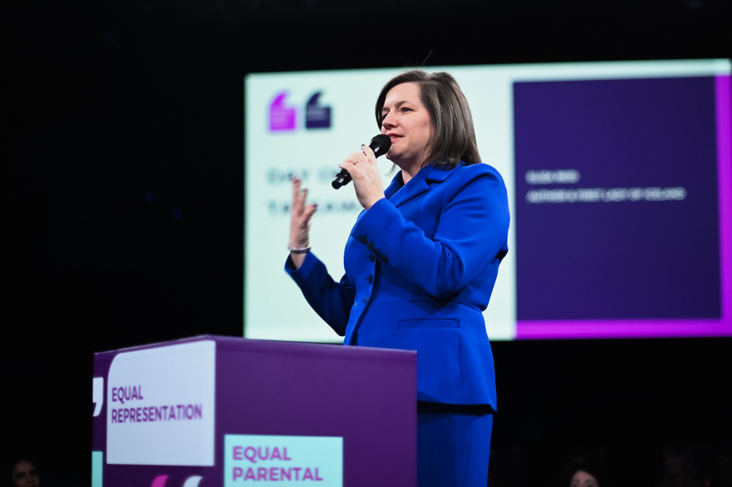 Eliza Reid speaking into a microphone on stage, wearing blue